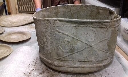 A large seemingly intact, slightly deformed lead tank sits on a table, it has embossed decoration in the pattern of an x in a rectangle with circles in each segment.