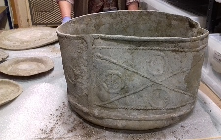A large seemingly intact, slightly deformed lead tank sits on a table, it has embossed decoration in the pattern of an x in a rectangle with circles in each segment.