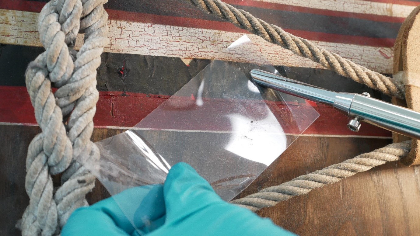 A gloved hand holding sheet of clear protective material under a heated spatula pressing onto the layer of black paint