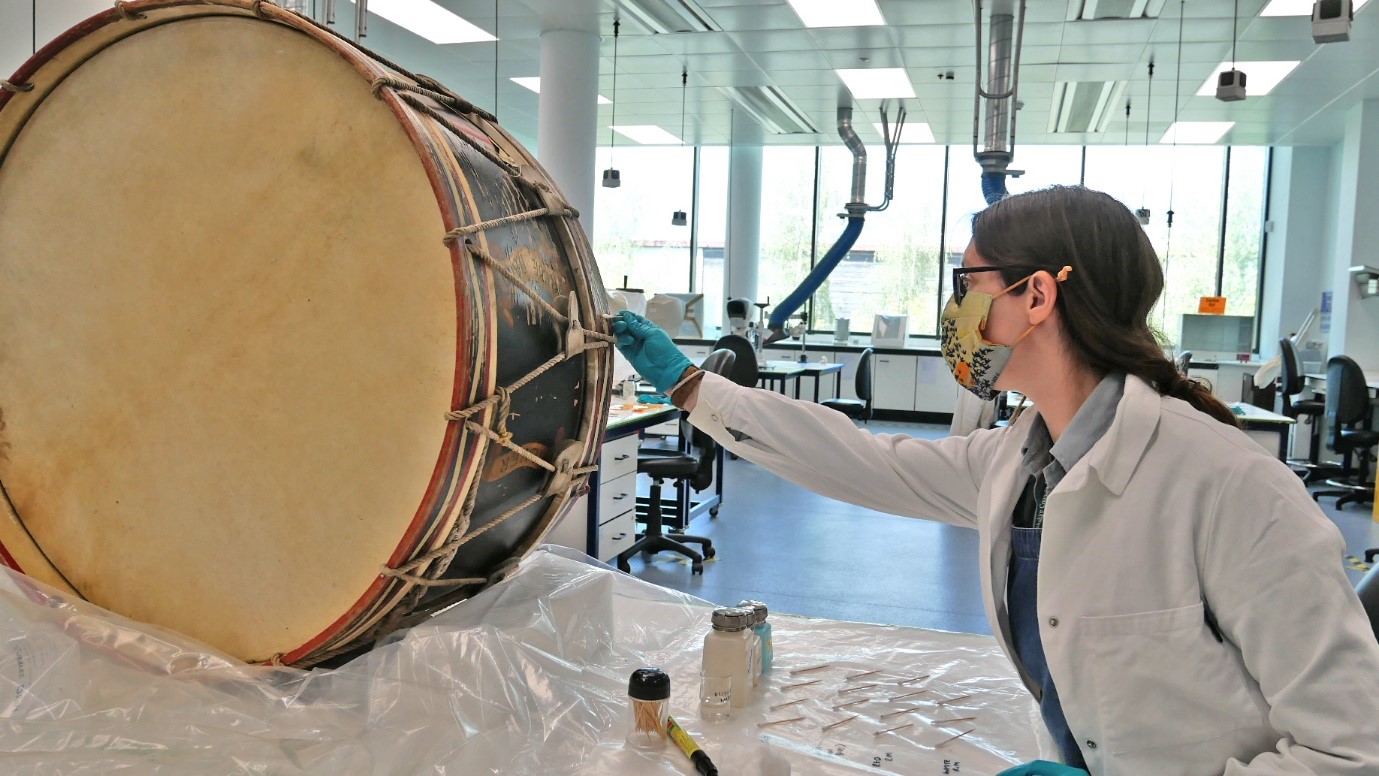 Image of the full drum with a conservator in a lab wearing gloves, white coat and a mask applying a solvent to the side of the drum with a cotton bud