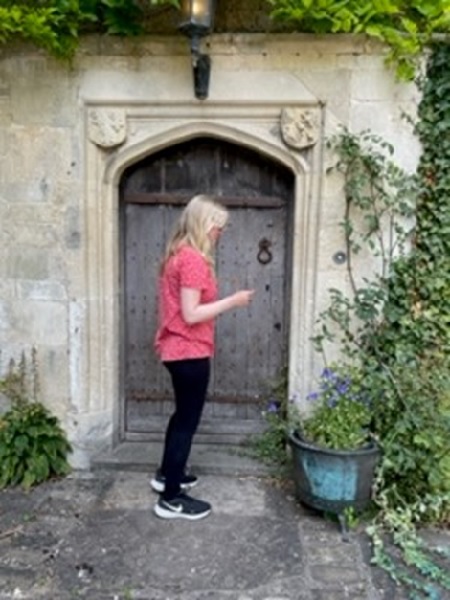 A girl in a red top, black trousers and blond hair is stood in front of an old wooden arched door surrounded by carved stonework and green plants.