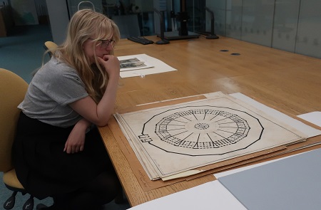 A girl in grey top sits with her chin resting on her fist sat at a table with a plan drawing of a polygonal building