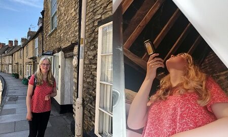 A teenage girl in red top, black trousers and blonde hair stands on the pavement next to a terrace of old buildings