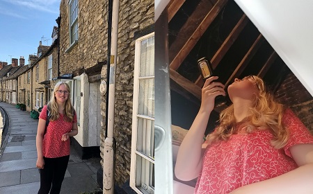 A teenage girl in red top, black trousers and blonde hair stands on the pavement next to a terrace of old buildings