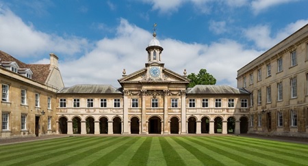 View of grand facade with open arcaded walk and clock tower, the wings of the building enclosing striped lawn.