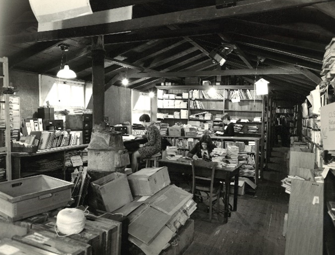 Interior of wooden hut lined with shelves, trolleys, three staff working and boxes piled in foreground.