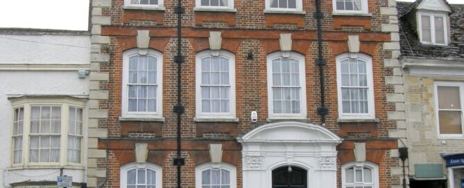 A three-storey red-brick facade with sash windows and front door with white arched pediment surround, standing taller than the buildings on either side.