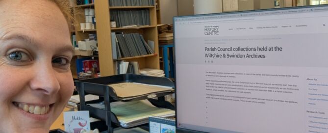 A thumbs-up selfie showing light-haired woman grinning at the camera with desktop computer showing webpage about parish council collections at WSHC behind.