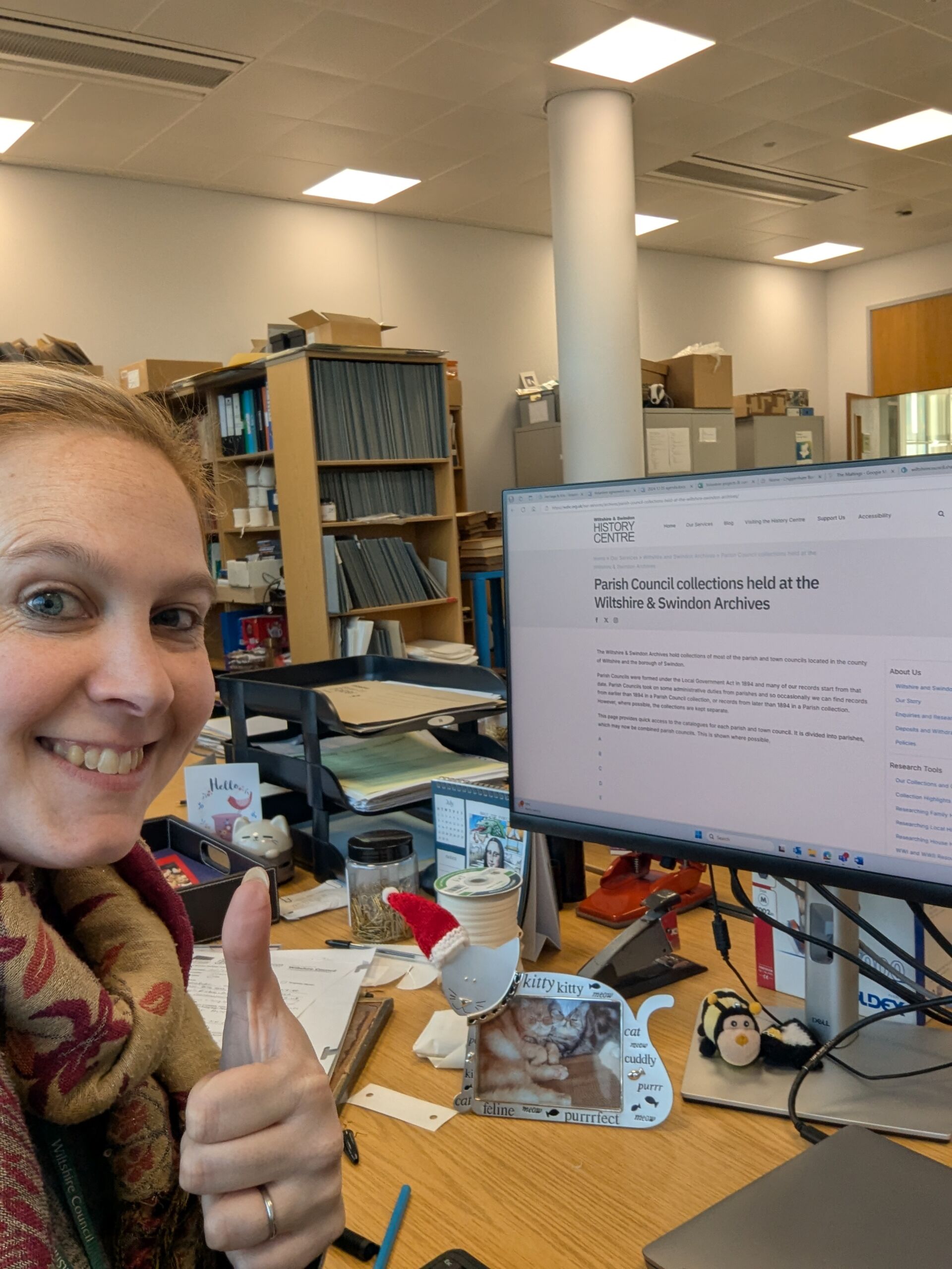 A thumbs-up selfie showing light-haired woman grinning at the camera with desktop computer showing webpage about parish council collections at WSHC behind.