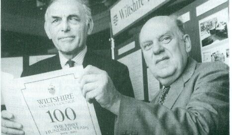 Black and white photograph showing open booklet titled "Wiltshire County Council: The First 100 years" being held by two men in suits.
