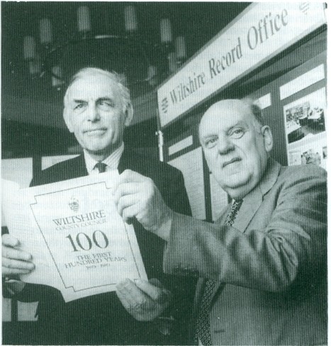 Black and white photograph showing open booklet titled "Wiltshire County Council: The First 100 years" being held by two men in suits.