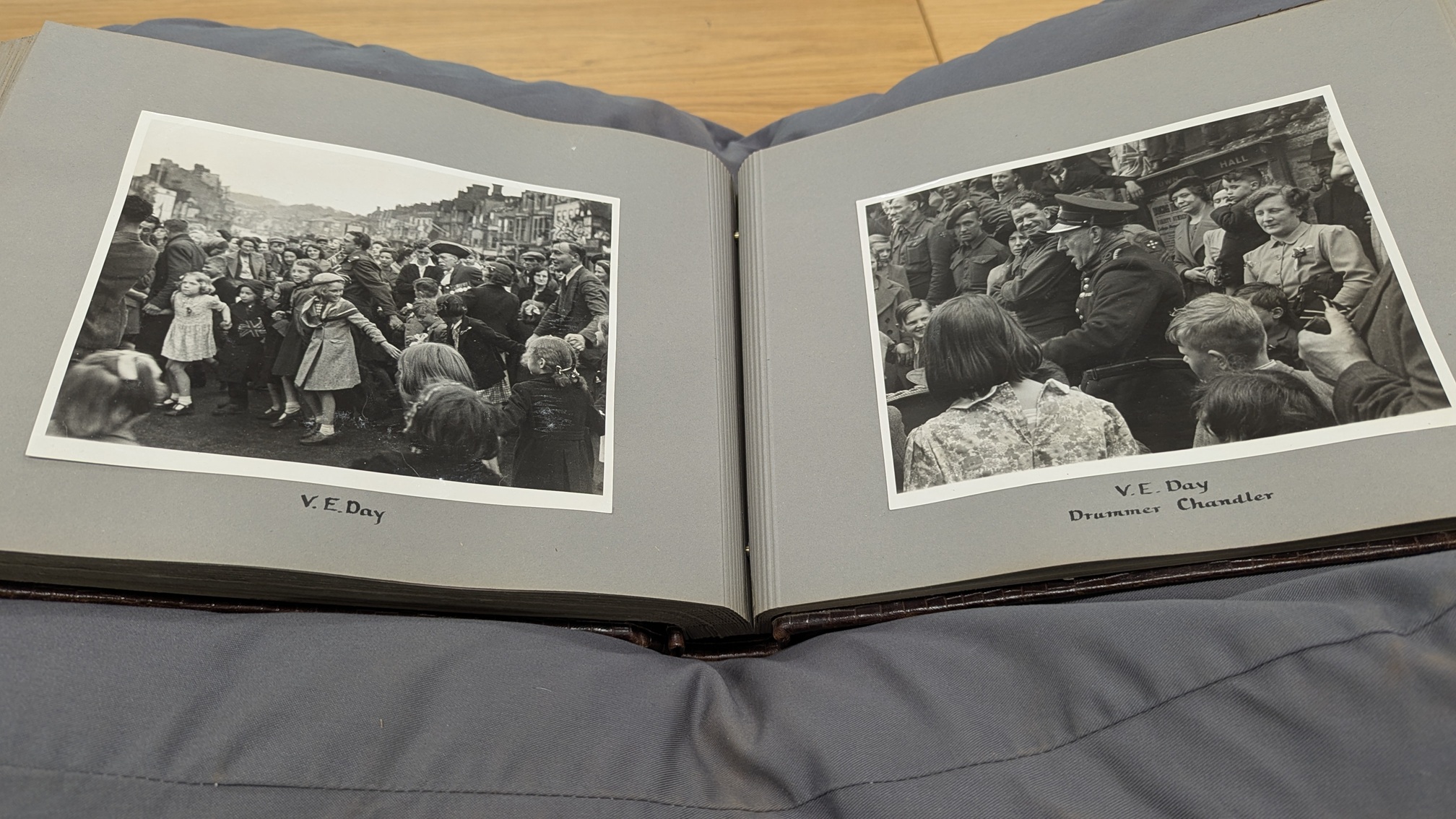 Two black and white photographs in album which is open and supported on a cushion. The photographs show a crowds of people.