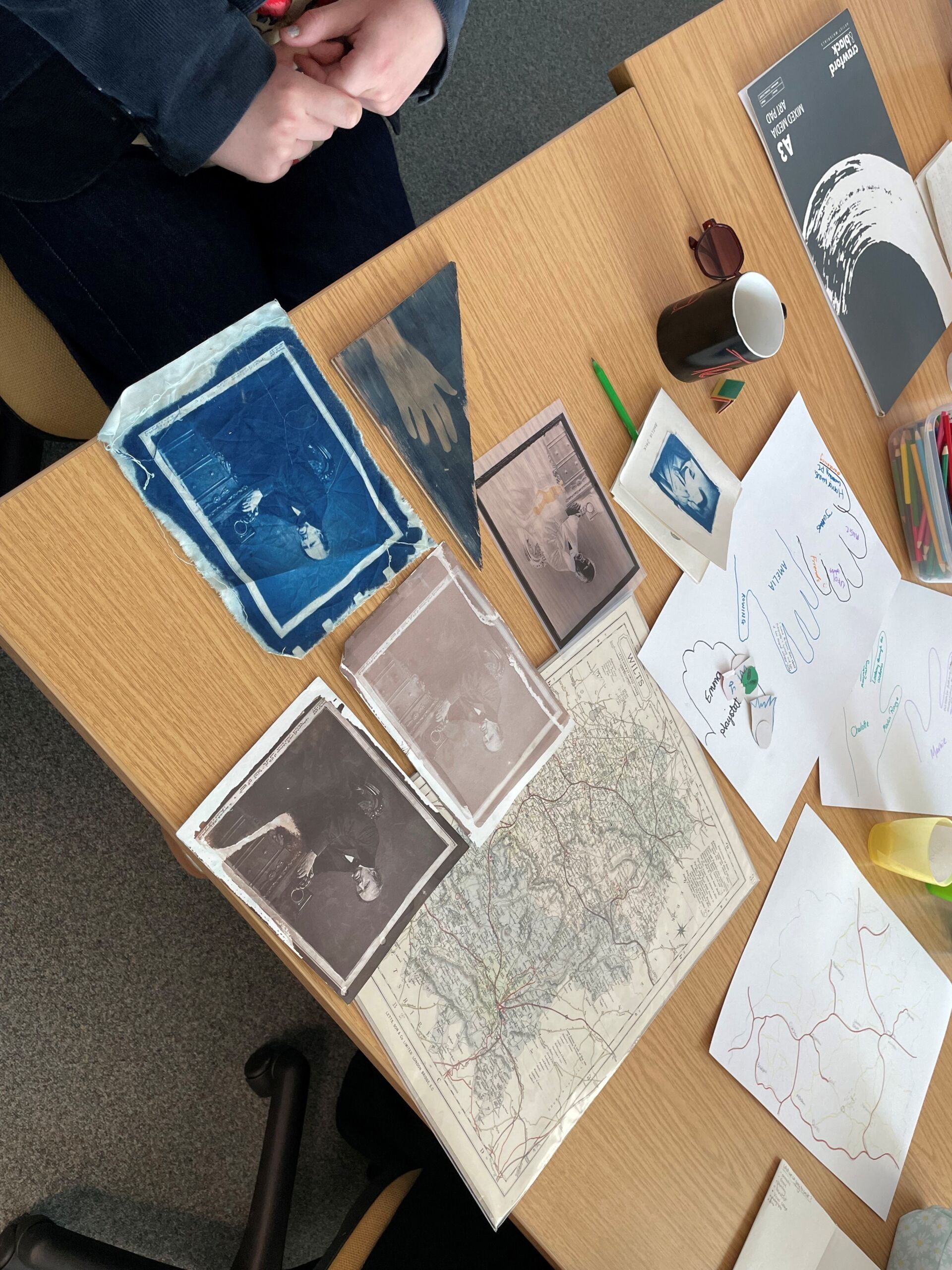 A table spread with different types of photographic development paper depicting a portrait, impression of a hand, drawings of hands, and a printed map.