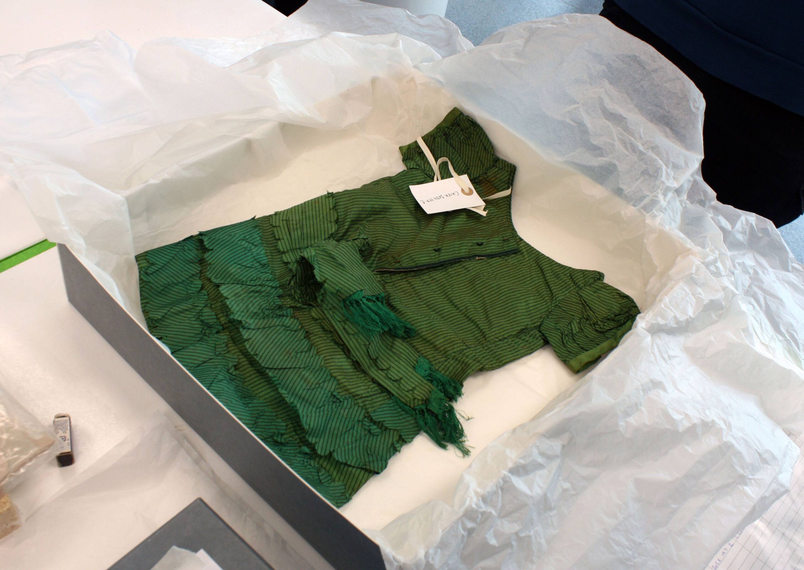 A small, vibrant darkish green dress with ruffles on the skirt and a white label laid on top left shoulder, lying on white tissue paper in a card storage box.