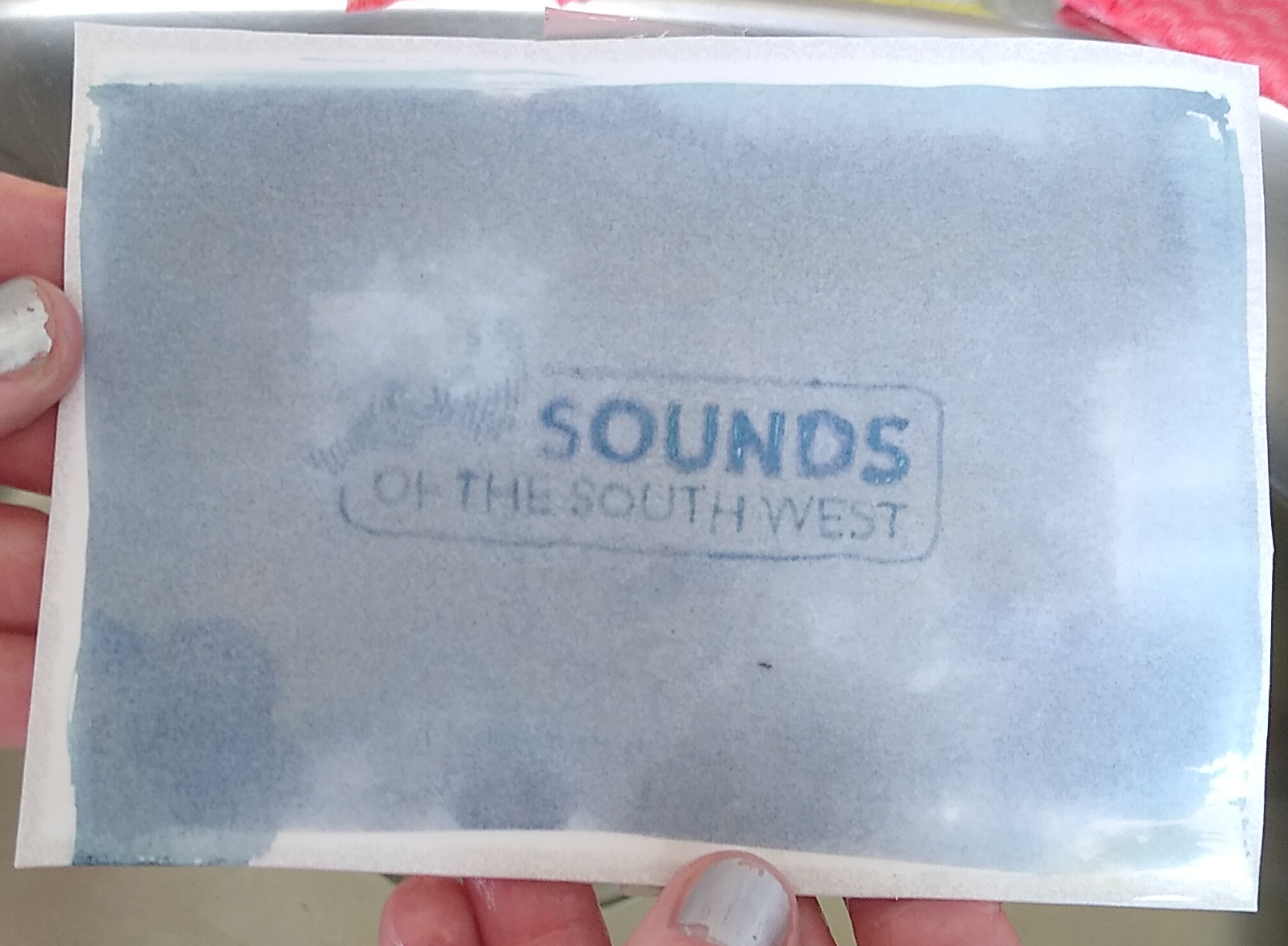 SoSW logo after washing – cropped An image of the Sounds of the South West logo emerging as a photographic print on a piece of paper which has been prepared using the calotype process, being held just after washing