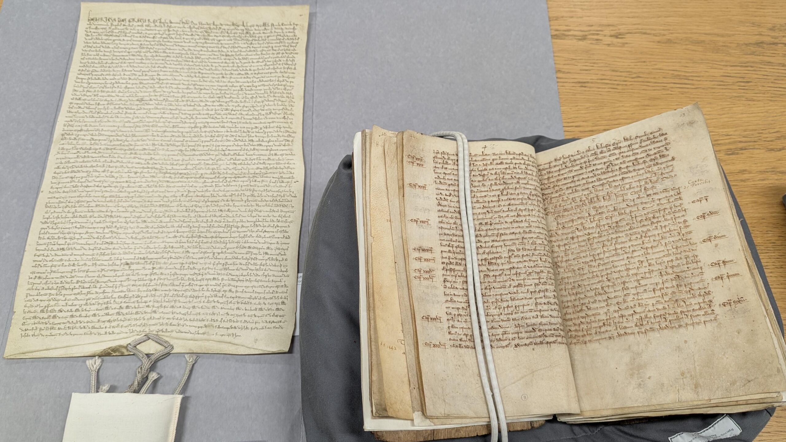 Open manuscript book with string weights to hold page open, supported on a cushion with large flat copy of manuscript document on the left with appended large seal contained within a canvas padded bag.