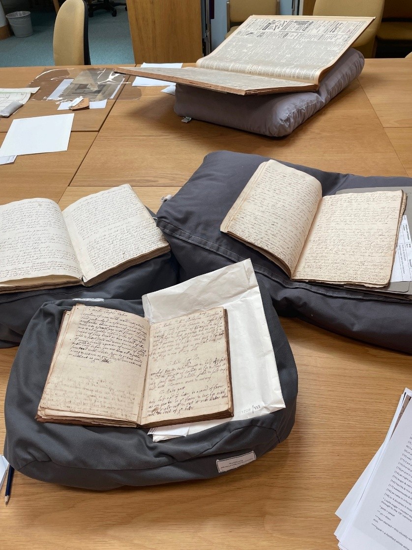 A table displaying several old books and manuscripts on cushioned supports. The documents are open to pages filled with dense handwriting