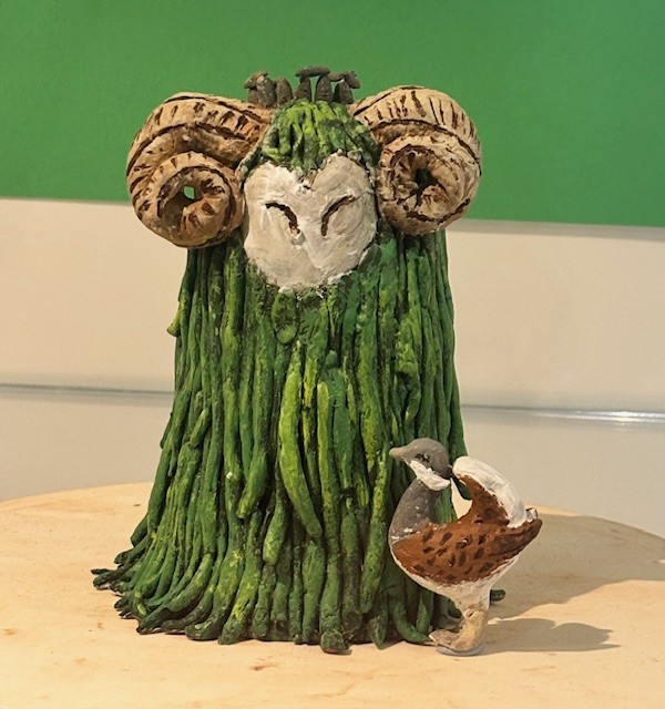 Sculpture of a fantastical creature with ram-like curled horns, an owl-like face, and a moss-textured green body, accompanied by a smaller bird sculpture in front, appearing to look up at the larger figure