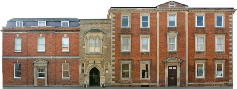 Historic red-brick building with Gothic-style stone archway entrance, featuring dormer windows on the left, ornate detailing in the center, and symmetrical three-story structure on the right