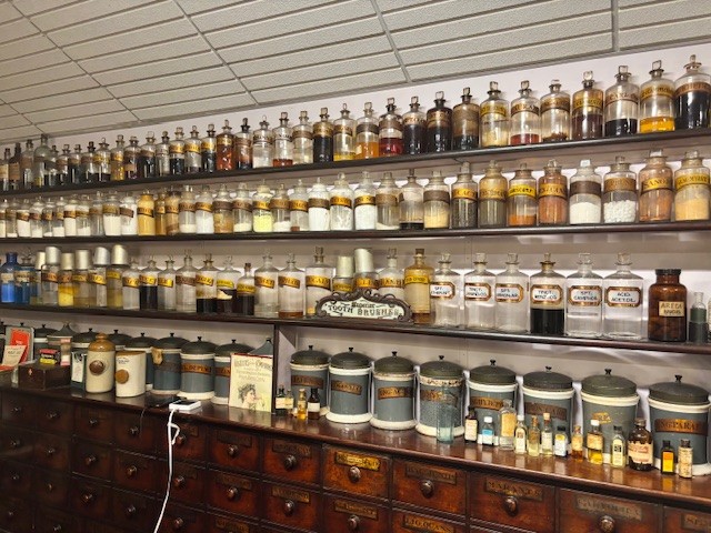 Vintage apothecary interior with shelves lined with labeled glass jars containing powders, liquids, and chemicals such as sulphur, magnesia, and acid; larger containers and drawers are visible below, reflecting historical pharmaceutical practices.