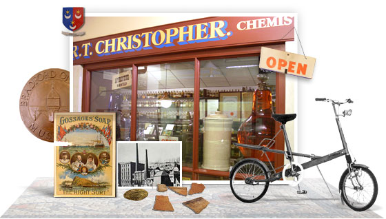 Storefront of 'R.T. Christopher Chemist' with an 'OPEN' sign on the door; inside are shelves and glass containers. In front, a display includes a 'BRADFORD ON AVON' coin, Gossage's Soap advertisement with three figures, a black-and-white photo of industrial buildings, pottery or bricks, and a bicycle—suggesting a historical exhibit of pharmacy and local heritage.