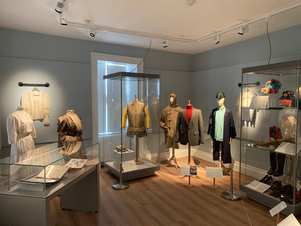 Museum exhibit room displaying historical clothing on mannequins and in glass cases, featuring garments such as a yellow-sleeved vest, brown coat, grey suit jacket with red accents, and various accessories like hats and shoes.