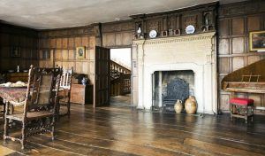 Historic room with wooden panelling and flooring, featuring a large ornate fireplace with ceramic pots and decorative plates above. A dining table with a patterned tablecloth and chairs sits to the left, while a piano with an upholstered stool is on the right. An open door in the background reveals a staircase with wooden railings.