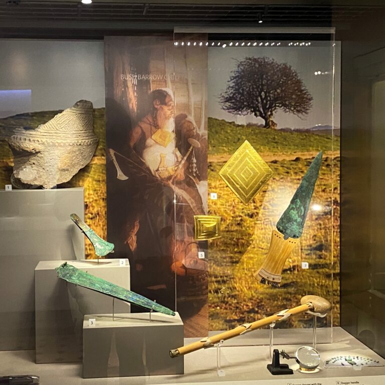 Museum display case featuring ancient artifacts including a large woven basket, greenish-blue metal tools or weapons, a decorated dagger, a long-handled object, and several gold-coloured geometric items. The backdrop includes an illustration of an ancient figure holding similar artifacts, set against a scenic landscape with a tree.