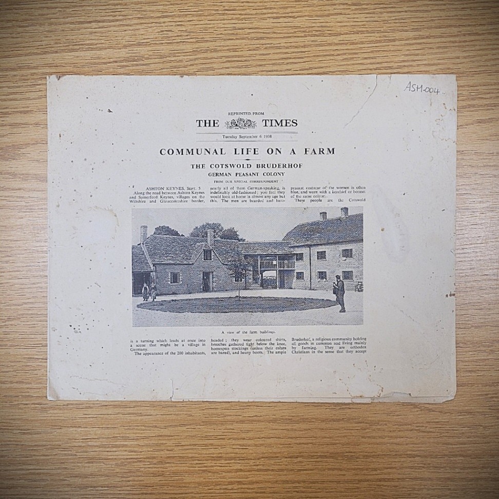 Printed text on off-white paper on wooden table titled 'The Times: Communal Life on a Farm' with small text and a large central image showing old stone building with round grass lawn in front and three figures stood and walking around the grass. 