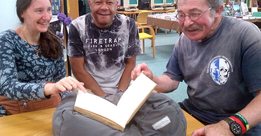 A group of three laughing people are sat with an open manuscript volume supported on a cushion on the table in front of them.