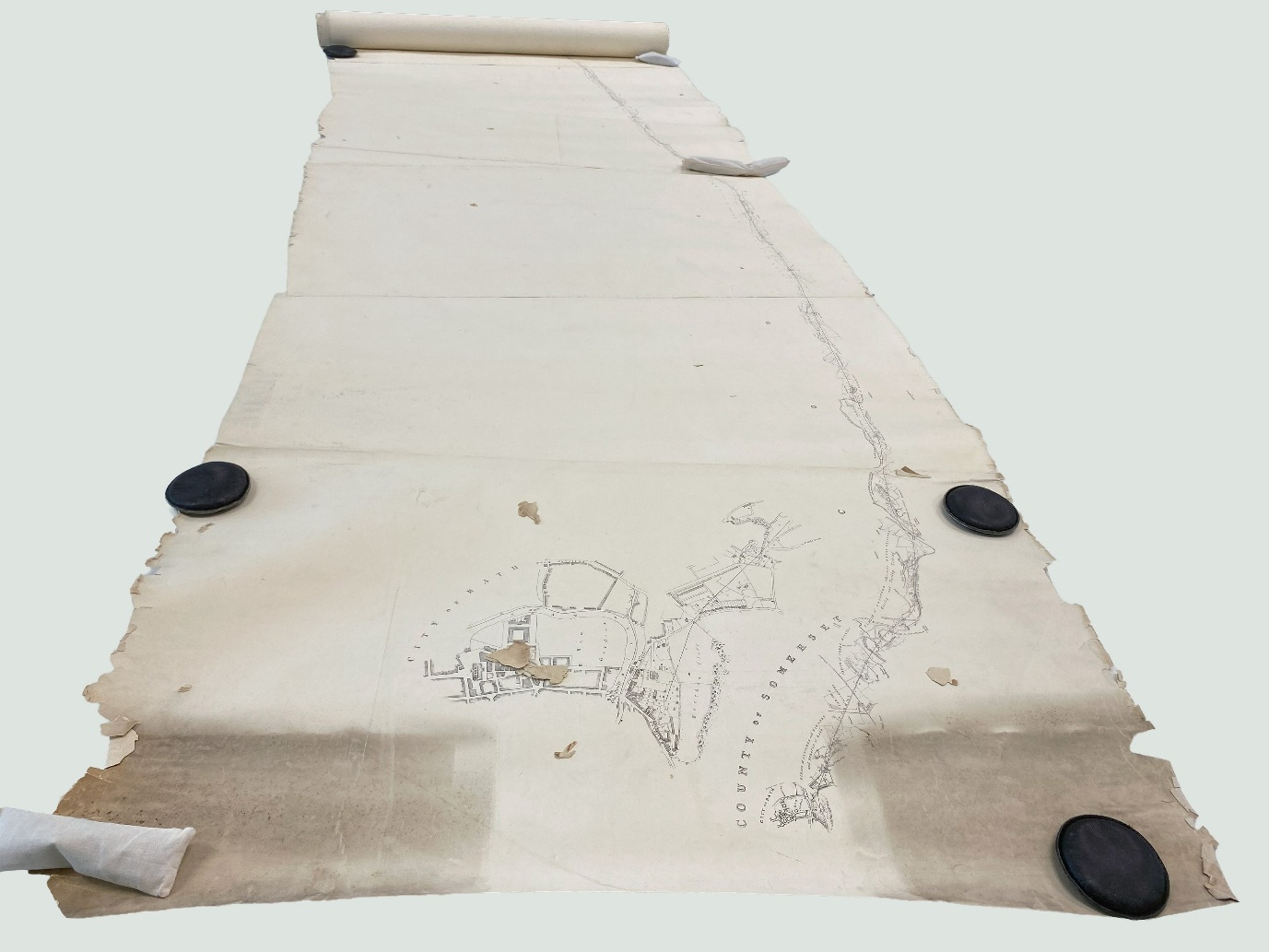 View along long rolled sheet held with weights showing fragments of paper on surface, and the linear route map stretching along the bottom of the sheets.