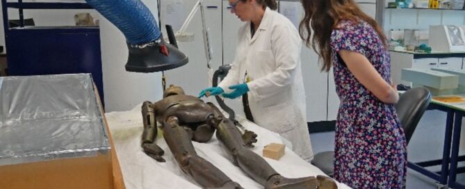 Conservator in white lab coat and blue latex gloves stood next to a table with lay figure laid flat on a white cloth, with a woman in floral dress stood alongside