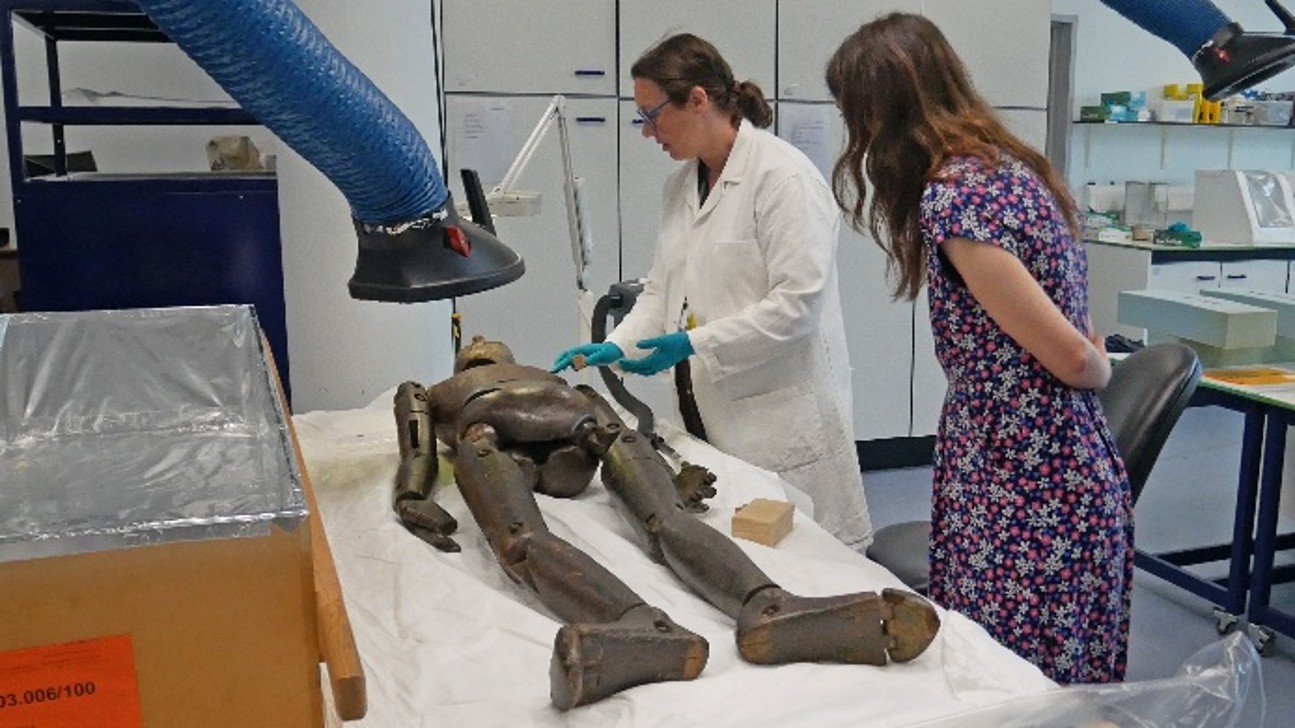 Conservator in white lab coat and blue latex gloves stood next to a table with lay figure laid flat on a white cloth, with a woman in floral dress stood alongside